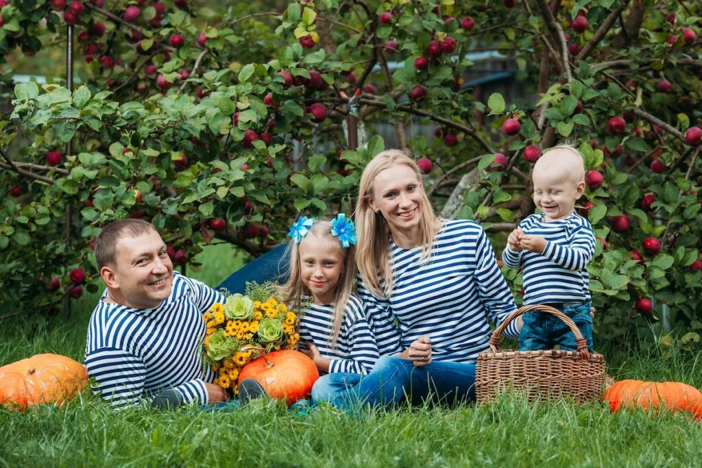 a person and two children sitting in grass by a tree with pumpkins