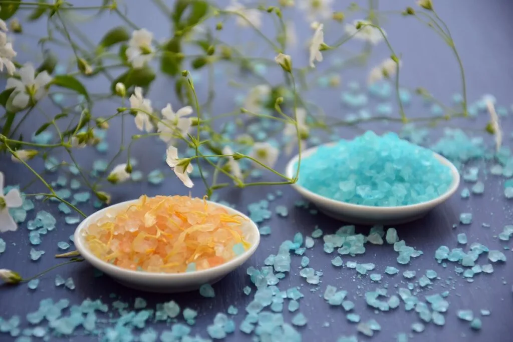 A close-up of colorful bath salts in dishes surrounded by delicate white flowers on a blue surface.