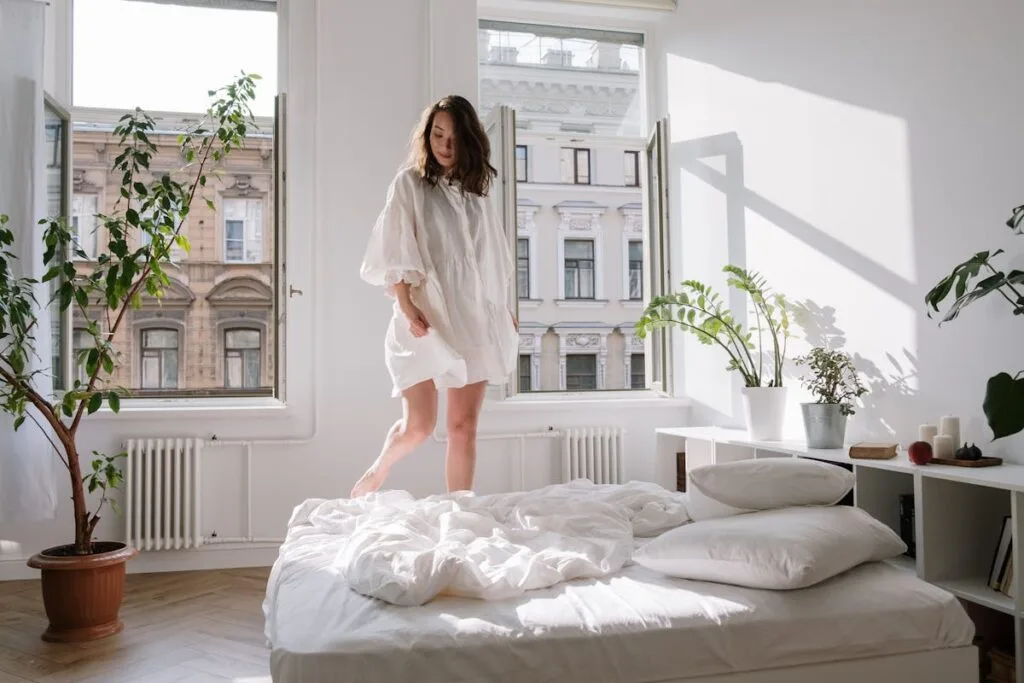 A woman enjoys a peaceful morning in a sunlit bedroom with open windows and potted plants.