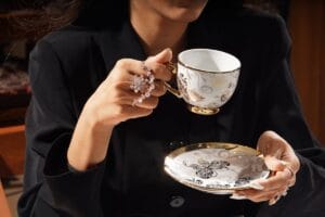 A close-up of a woman in black attire holding a beautifully decorated teacup with matching saucer.